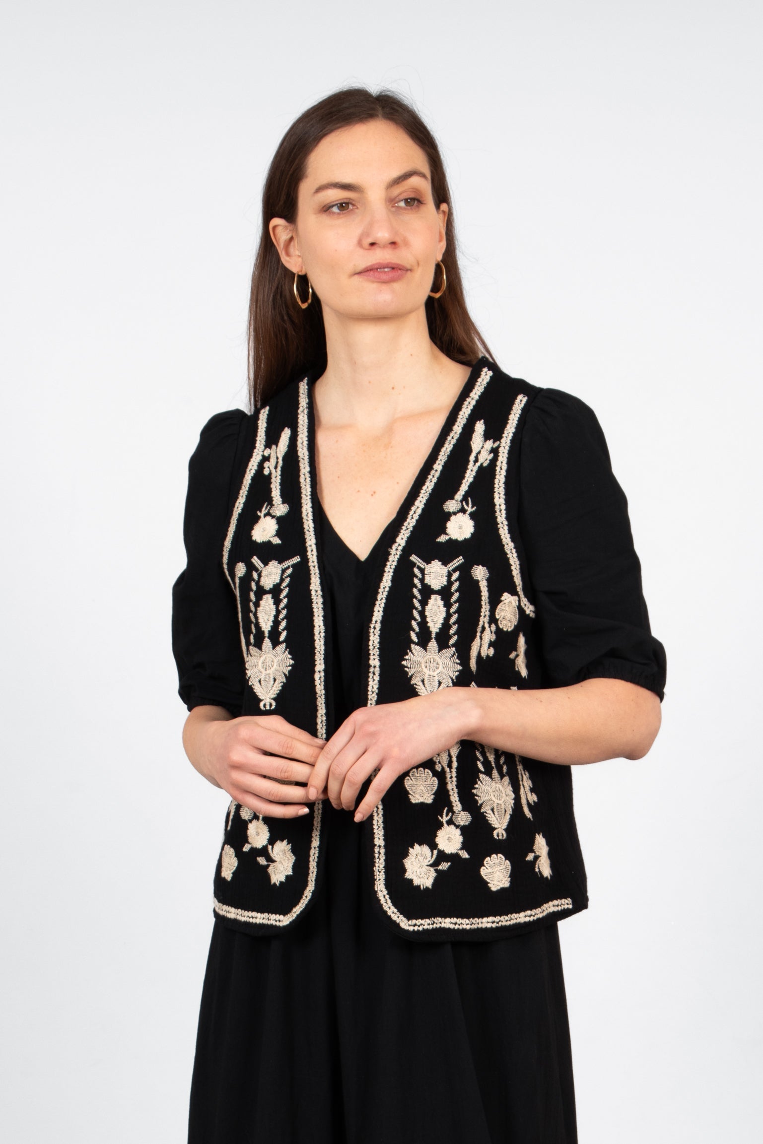 model wearing a black sleeveless lightweight waistcoat. the pattern on the waistcoat is an embroidered vintage floral design in ecru.
