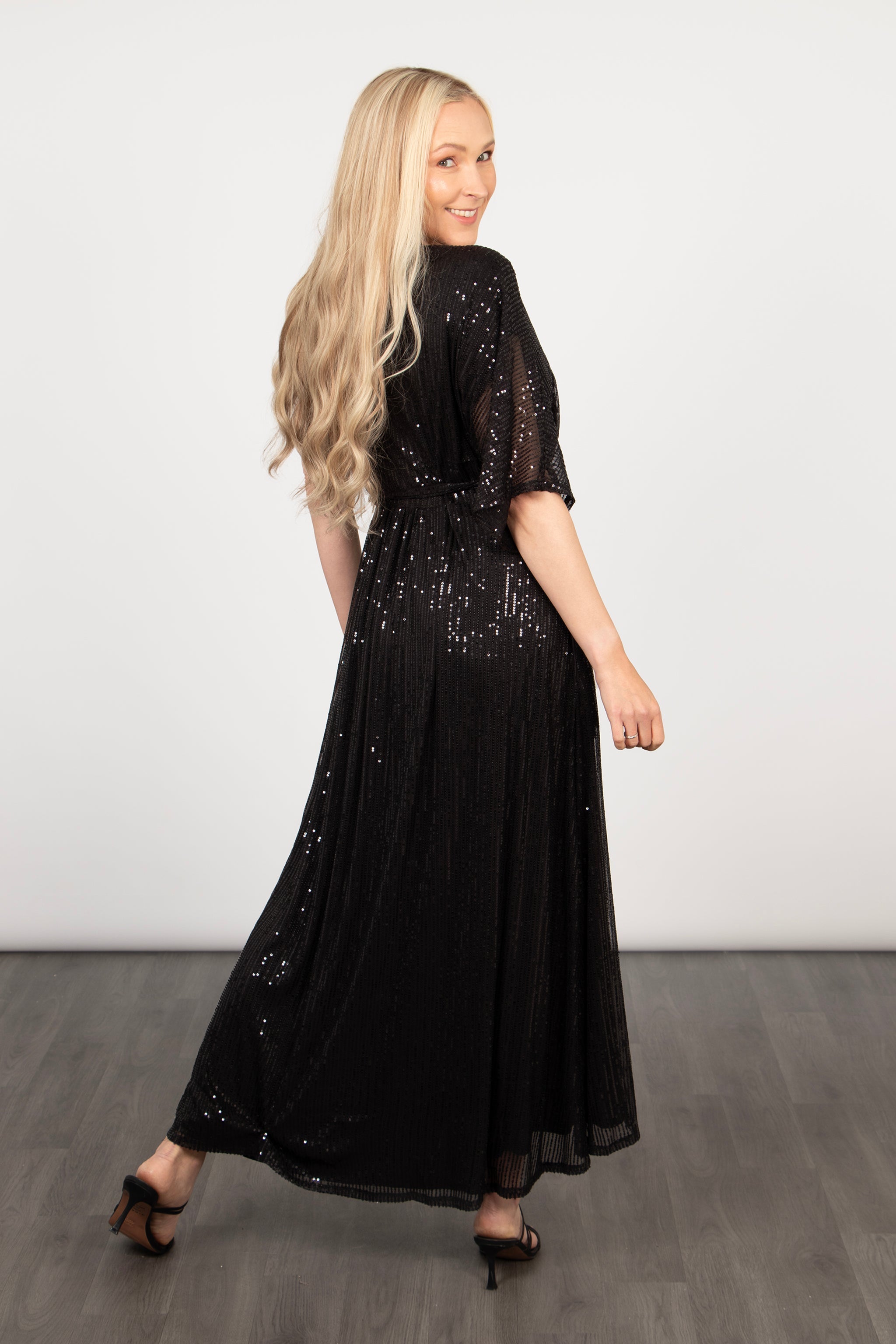 model showing the back of the black sequin wrap dress. the hem is longer at the back than at the front, it is maxi length at the back