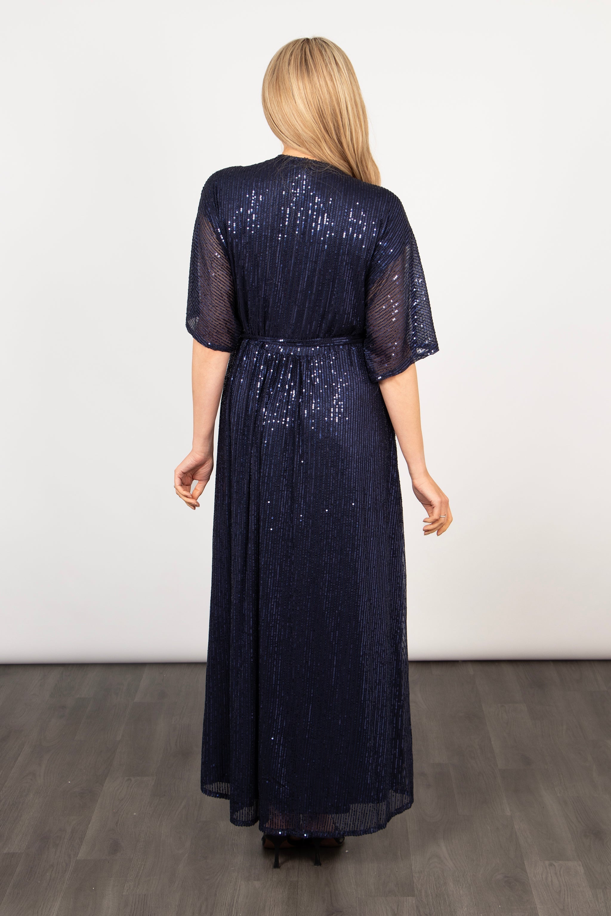 model showing the back of the navy blue sequin wrap dress, showing the maxi length hem at the back, all over sequin sparkle and waist tie